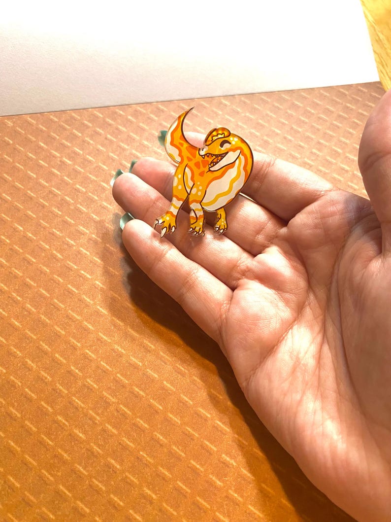 May include: A yellow and orange enamel pin of a cartoon dinosaur with a toothy grin. The dinosaur is standing on its hind legs with its tail curled up.