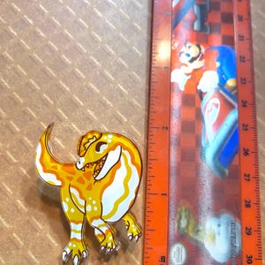 May include: A yellow and white cartoon dinosaur pin with a smiling face. The pin is about 2 inches tall and is made of clear acrylic.