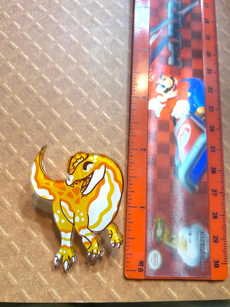 May include: A yellow and white cartoon dinosaur pin with a smiling face. The pin is about 2 inches tall and is made of clear acrylic.