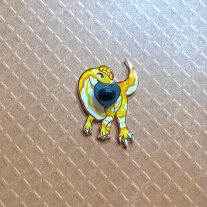 May include: A yellow and white enamel pin of a cartoon gecko with a black heart on its chest.