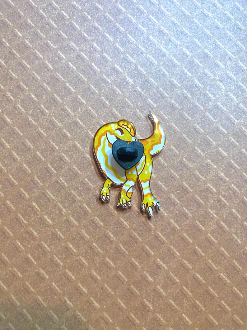 May include: A yellow and white enamel pin of a cartoon gecko with a black heart on its chest.