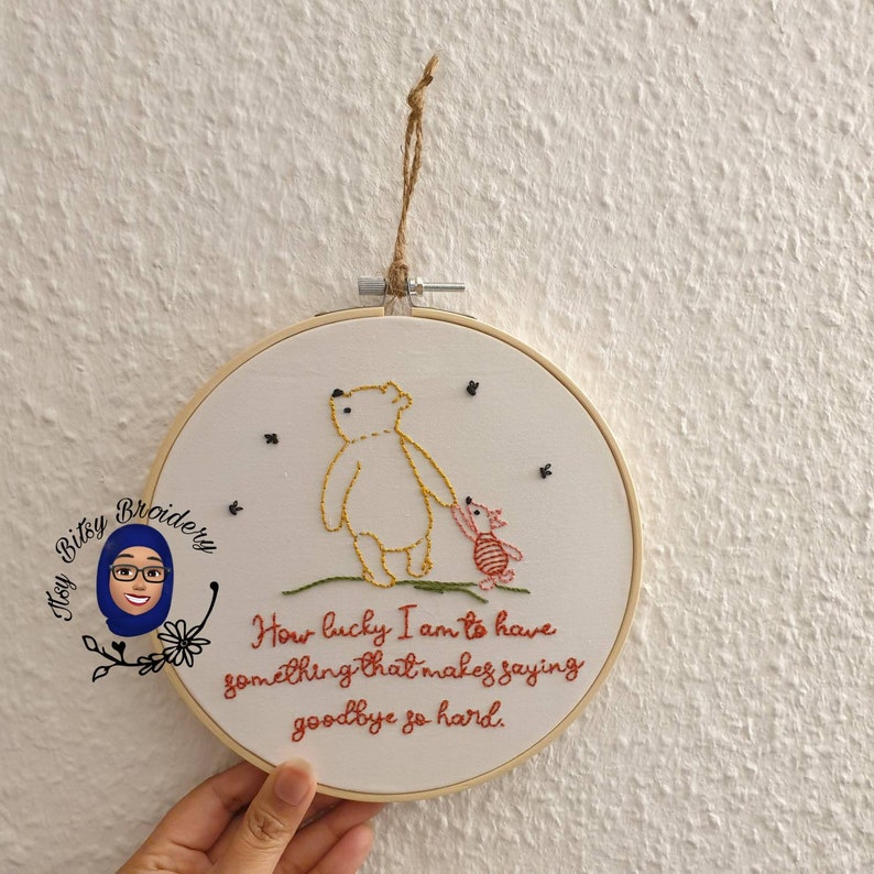 May include: Embroidered hoop art depicting Winnie the Pooh and Piglet, holding hands. The artwork is stitched with yellow, red, and pink thread on white fabric. The quote "How lucky I am to have something that makes saying goodbye so hard" is also embroidered.