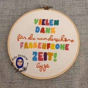 May include: Embroidered hoop art with the text "VIELEN DANK f&uuml;r die wundersch&ouml;ne FARBENFROHE ZEIT! Ayza" in colourful thread. The design includes a cartoon image of a woman in a hijab. The hoop is made of wood.