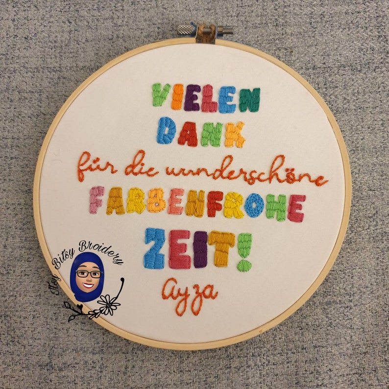 May include: Embroidered hoop art with the text "VIELEN DANK f&uuml;r die wundersch&ouml;ne FARBENFROHE ZEIT! Ayza" in colourful thread. The design includes a cartoon image of a woman in a hijab. The hoop is made of wood.