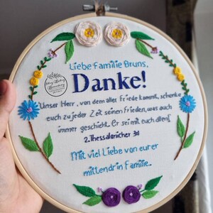 May include: Embroidered hoop art with the text "Liebe Familie Bruns, Danke!" and a Bible verse. The white fabric is decorated with blue, yellow, and purple floral embroidery. The wooden hoop is held by a hand.