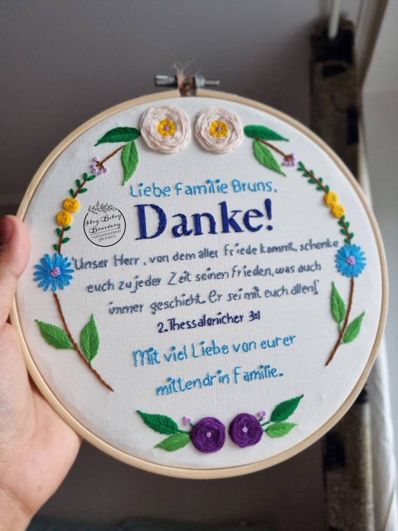 May include: Embroidered hoop art with the text "Liebe Familie Bruns, Danke!" and a Bible verse. The white fabric is decorated with blue, yellow, and purple floral embroidery. The wooden hoop is held by a hand.