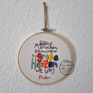 May include: Embroidered hoop art with the German text "Kleine Menschen brauchen so gro&szlig;e Herzen wie deins Milou." The text is in black, with the word "Herzen" in colorful letters. The hoop is suspended by a natural twine.