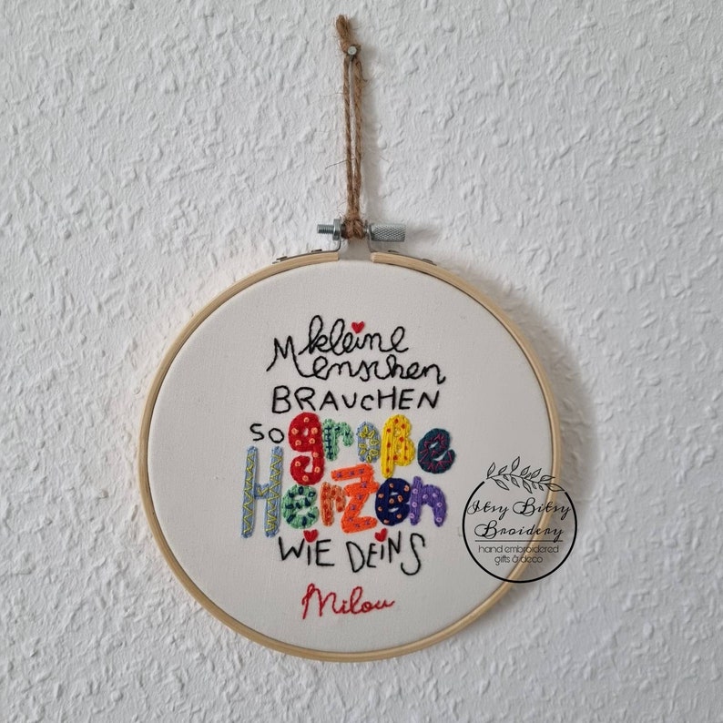 May include: Embroidered hoop art with the German text "Kleine Menschen brauchen so gro&szlig;e Herzen wie deins Milou." The text is in black, with the word "Herzen" in colorful letters. The hoop is suspended by a natural twine.