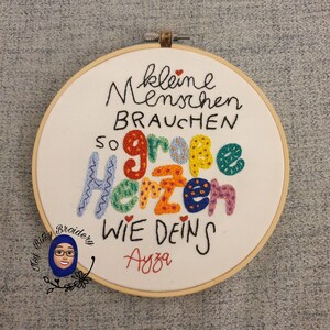 May include: Embroidered hoop art with the German text "kleine Menschen brauchen so gro&szlig;e Herzen wie deins" in various colours. The hoop is made of wood and the text is stitched on white fabric. The name "Ayza" is stitched in red.