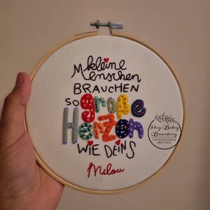 May include: Embroidered hoop art with the German text "Kleine Menschen brauchen so gro&szlig;e Herzen wie deins Milou." The colorful text is stitched on white fabric, framed by a wooden hoop. The design includes a small logo.