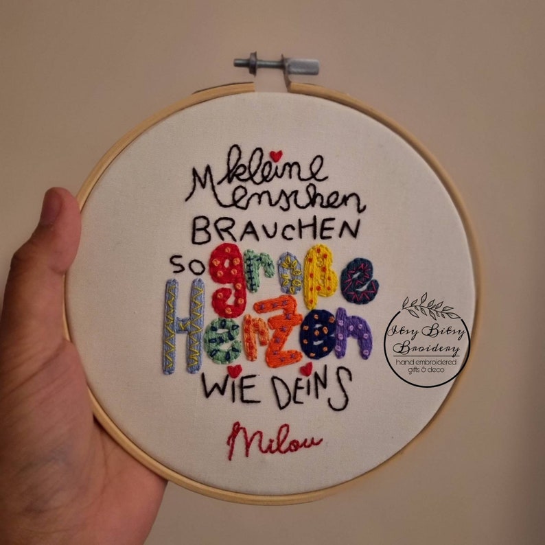 May include: Embroidered hoop art with the German text "Kleine Menschen brauchen so gro&szlig;e Herzen wie deins Milou." The colorful text is stitched on white fabric, framed by a wooden hoop. The design includes a small logo.