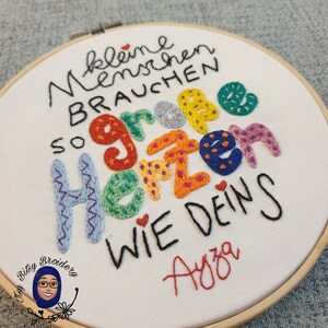 May include: Embroidered hoop art with the German text "kleine menschen brauchen so gro&szlig;e Herzen wie deins" in black and colourful thread. The artist's signature, "Ayza", is in red. The hoop is made of wood.