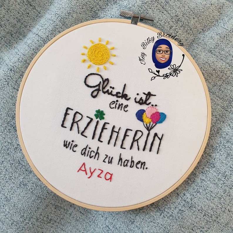 May include: Embroidered hoop art with the text "Gl&uuml;ck ist eine Erzieherin wie dich zu haben" and the name "Ayza". Features a yellow sun, a green four-leaf clover, and colourful balloons.