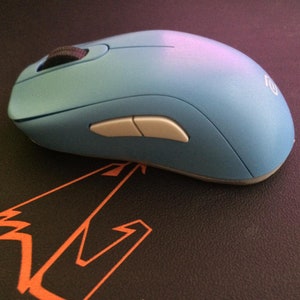 Könnte beinhalten: Eine hellblaue kabellose Computermaus mit zwei weißen Tasten auf der Oberseite. Die Maus befindet sich auf einer schwarzen Oberfläche mit einem orangefarbenen Design.