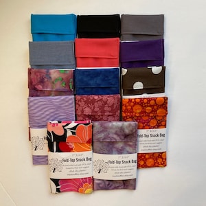 May include: A collection of 12 reusable snack bags made from fabric. The bags are in various colors and patterns, including floral, polka dot, and solid colors. The bags are 7 inches by 5.5 inches and have a fold-top closure. The bags are labeled "Fold-Top Snack Bag" and have the text "lined with food-safe PUL, great for fresh and veggie snacks, ditch the plastic, madstuffbiz.etsy.com".