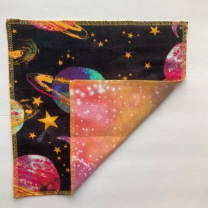 May include: A black fabric square with a colorful space-themed print featuring planets, stars, and a galaxy design. The fabric is folded in half to reveal a pink, orange, and yellow galaxy print on the other side.