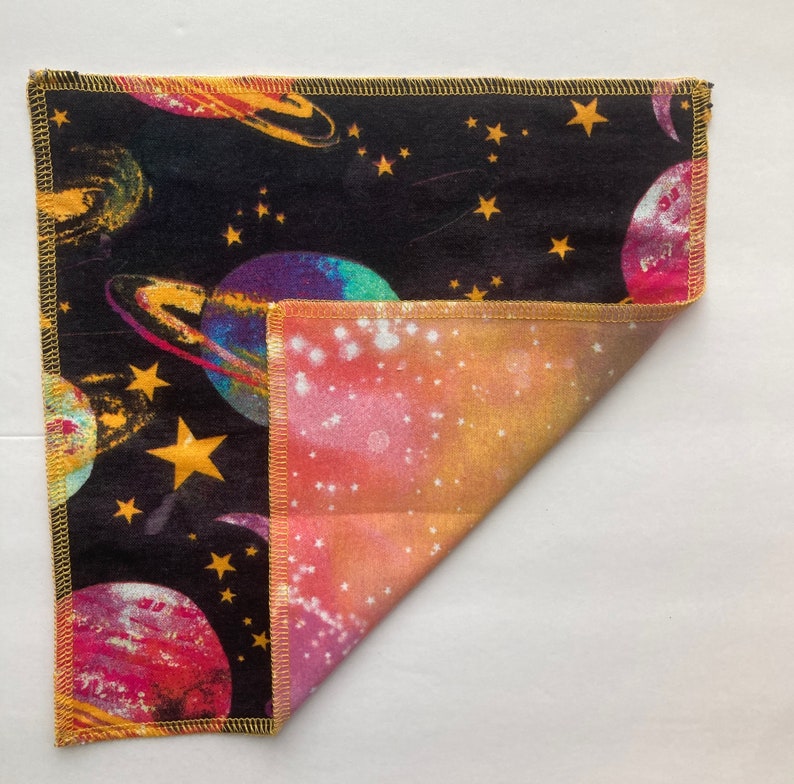 May include: A black fabric square with a colorful space-themed print featuring planets, stars, and a galaxy design. The fabric is folded in half to reveal a pink, orange, and yellow galaxy print on the other side.