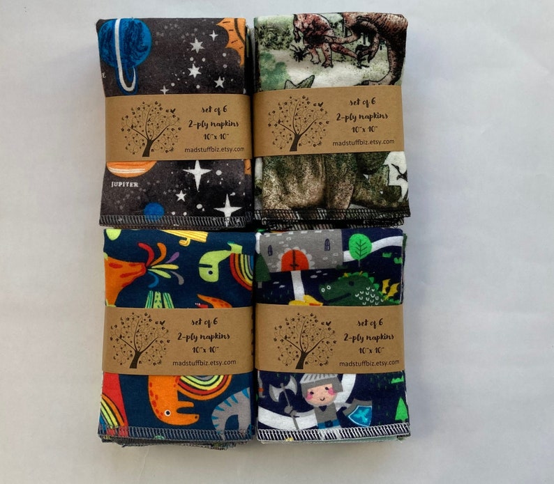 May include: Set of six 2-ply cloth napkins with various colorful prints. The prints include a space theme, dinosaurs, a knight, and a dragon. Each napkin measures 10 inches by 10 inches.