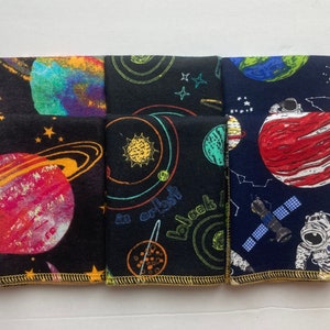 May include: Five fabric squares with a space theme. The squares are black with colorful planets, stars, and spacecrafts. One square is blue with a red planet, a spaceship, and an astronaut. The squares have a gold trim.