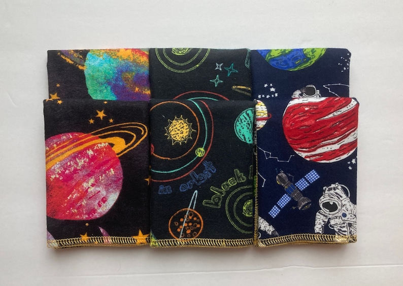 May include: Five fabric squares with a space theme. The squares are black with colorful planets, stars, and spacecrafts. One square is blue with a red planet, a spaceship, and an astronaut. The squares have a gold trim.