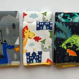 May include: Three fabric squares with a dragon theme. The first square is gray with a blue dragon breathing fire, the second is white with a yellow dragon and a blue castle, and the third is black with a green dragon and stars.
