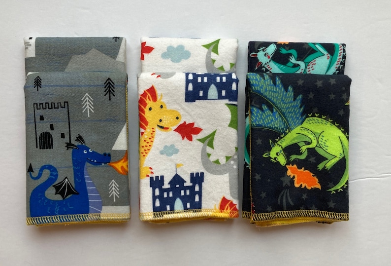 May include: Three fabric squares with a dragon theme. The first square is gray with a blue dragon breathing fire, the second is white with a yellow dragon and a blue castle, and the third is black with a green dragon and stars.