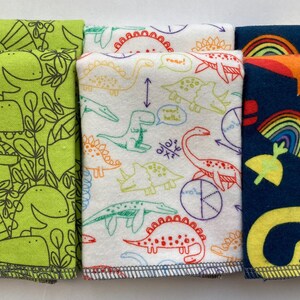 May include: Three fabric squares with dinosaur designs. The first square is green with black outlines of dinosaurs. The second square is white with colorful outlines of dinosaurs. The third square is dark blue with yellow dinosaurs and rainbows.