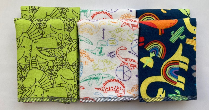 May include: Three fabric squares with dinosaur designs. The first square is green with black outlines of dinosaurs. The second square is white with colorful outlines of dinosaurs. The third square is dark blue with yellow dinosaurs and rainbows.