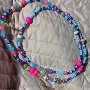 May include: A beaded necklace featuring a variety of colorful beads in shades of light blue, purple, pink, and white. The strand includes round, translucent, opaque, and pearl-like beads, along with a single white bead decorated with a black floral pattern.