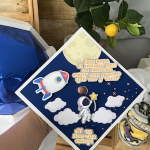 Op de afbeelding: Een afstudeerhoed met een ruimtethema. De hoed heeft een blauwe achtergrond met een raket, astronaut, wolken, maan en sterren. De Spaanse tekst luidt "Que sepa el mundo que en marcha estoy con mucho que ver y vivir" en "Si en marcha estoy!"