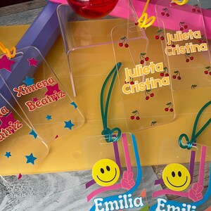 Puede incluir: Etiquetas de nombre de acrílico transparente con diseños coloridos. Algunas etiquetas presentan nombres como "Ximena Beatriz" y "Julieta Cristina" con gráficos de estrellas y cerezas. Otras tienen caras sonrientes y el nombre "Emilia Rosa".