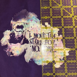 May include: A purple fabric with a graphic of a person and the text "I MOVE THE STARS FOR NO ONE" in white. The graphic is over a colorful, watercolor-like design. A yellow ruler is on the right side of the image.