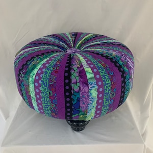 May include: A purple and green patterned fabric ottoman with a black base. The ottoman is round and has a tufted top.