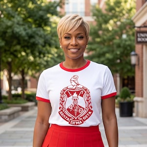 May include: A white t-shirt with red trim on the sleeves and neckline, featuring a red Delta Sigma Theta crest graphic on the chest, paired with a solid red pleated skirt. The crest includes a torch, laurel leaves, and Greek lettering.