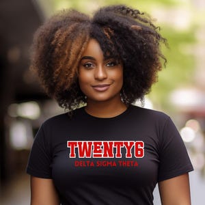 May include: Black t-shirt with the text "TWENTY6" in red block letters above "DELTA SIGMA THETA" in red. The shirt is paired with a camouflage print skirt and a black belt. The image is taken outdoors.