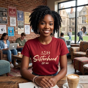 May include: A woman wearing a maroon t-shirt with the text "ΔΣΘ Sisterhood Est. 1913" in a coffee shop setting. The image includes a coffee cup, a notebook, and posters on the brick wall. Other people are in the background.