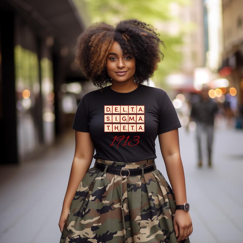 Puede incluir: Una camiseta negra con el texto "DELTA SIGMA THETA 1913" en rojo y blanco. La persona lleva una falda de camuflaje y un cintur&oacute;n negro. Un reloj est&aacute; en la mu&ntilde;eca.