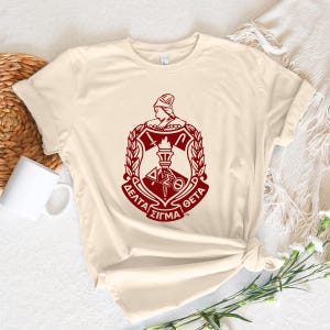 May include: A cream colored t-shirt with a red Greek sorority crest featuring a woman's head, a torch, and the letters "Delta Sigma Theta".
