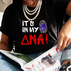 May include: Black t-shirt with the text "IT'S IN MY ΔNA!" in white and red. A fingerprint graphic with a purple flower is on the right side of the shirt. The person is wearing a pearl necklace and reading a magazine.