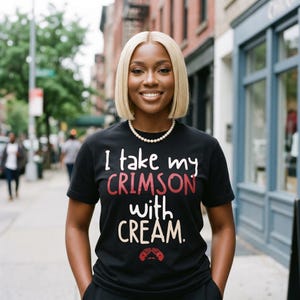 May include: Black t-shirt with the text "I take my CRIMSON with CREAM." in white and red lettering. The shirt features a graphic of two hands. The person is wearing a pearl necklace and has blonde hair.