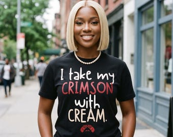 May include: Black t-shirt with the text "I take my CRIMSON with CREAM." in white and red lettering. The shirt features a graphic of two hands. The person is wearing a pearl necklace and has blonde hair.