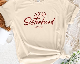 Puede incluir: Una camiseta de color crema con un gráfico rojo que dice "ΔΣΘ Sisterhood est. 1913".