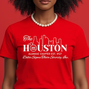 May include: A bright red short-sleeved t-shirt featuring a white graphic of the Houston skyline above the text The HOUSTON 100 Years ALUMNAE CHAPTER EST. 1927 Delta Sigma Theta Sorority, Inc. The shirt is worn with a white beaded necklace.