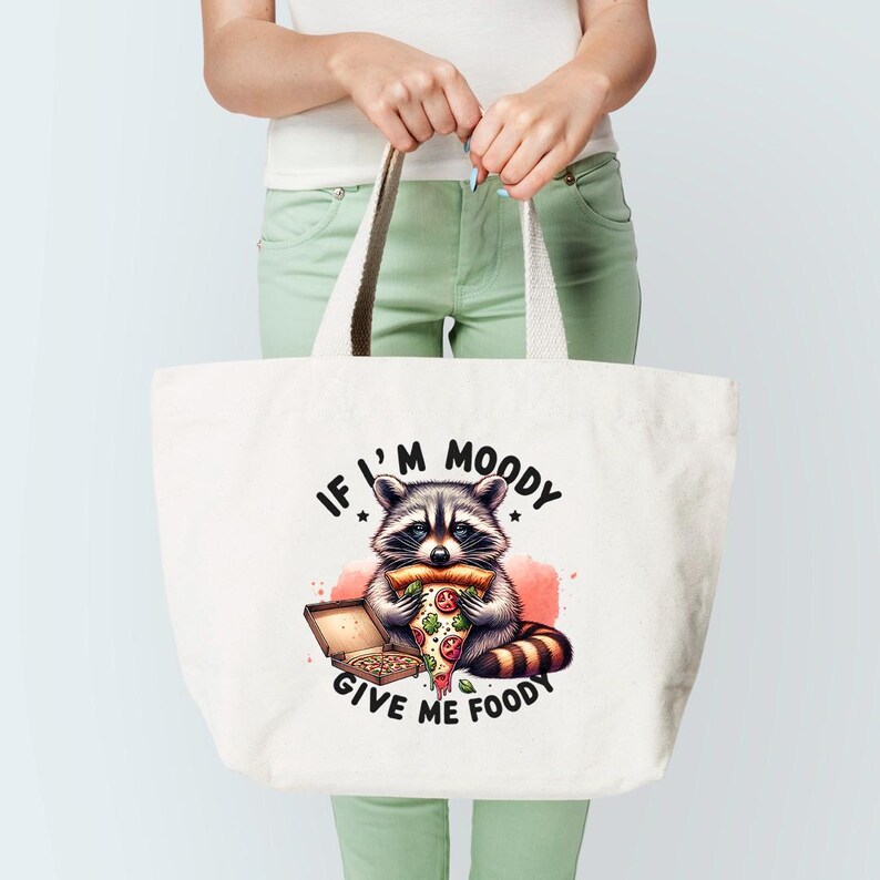 May include: White canvas tote bag with a cartoon raccoon eating a pizza slice. The text on the bag reads "If I'm Moody Give Me Foody".