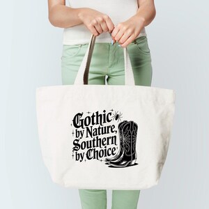 Puede incluir: Un bolso tote blanco con el texto "Gothic by Nature, Southern by Choice" en negro. Un gr&aacute;fico de bota de vaquero negra est&aacute; en el lado derecho. El bolso tiene asas color crema.