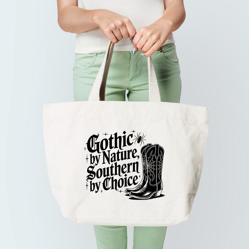 Puede incluir: Un bolso tote blanco con el texto "Gothic by Nature, Southern by Choice" en negro. Un gr&aacute;fico de bota de vaquero negra est&aacute; en el lado derecho. El bolso tiene asas color crema.