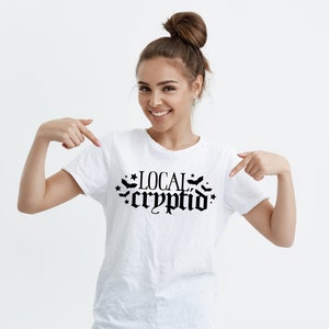 May include: White t-shirt with black text that reads "LOCAL cryptid" with bats and stars around the text.
