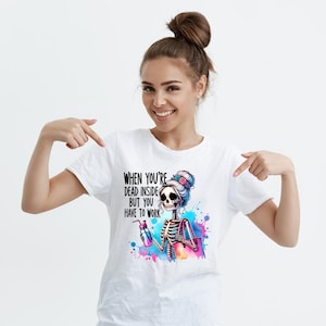 May include: White t-shirt with a colorful graphic of a skeleton with a bun in her hair holding a water bottle and a shopping bag. The text on the shirt reads "When you're dead inside but you have to work."