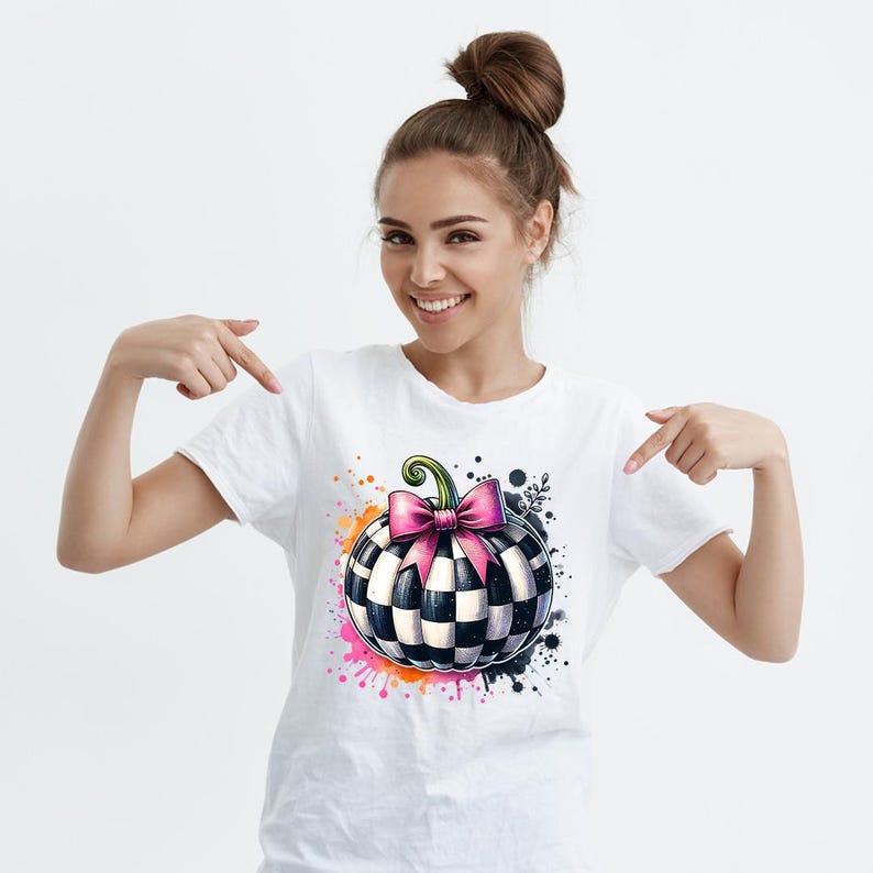 May include: White t-shirt featuring a watercolor design of a checkered pumpkin with a pink bow and green stem. The pumpkin is surrounded by colorful paint splatters. A fun and festive graphic tee.
