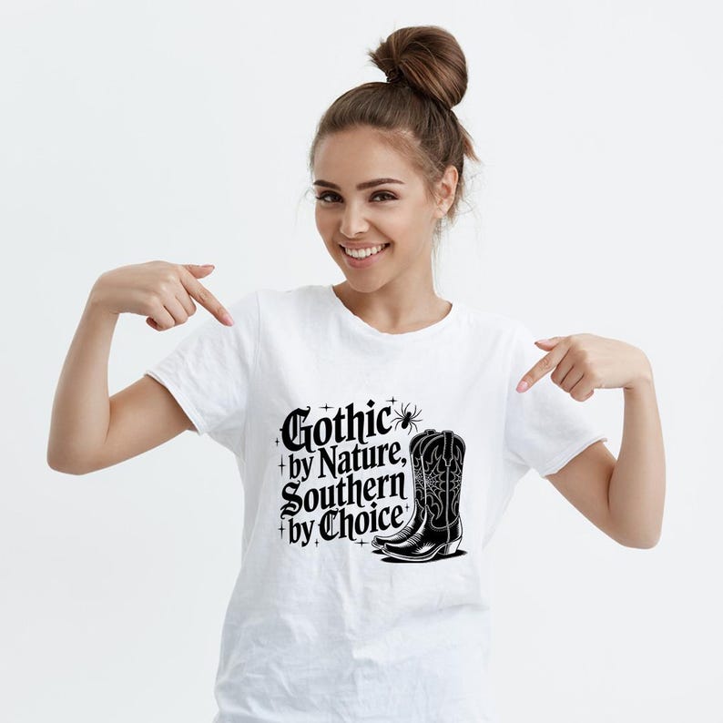 Puede incluir: Camiseta blanca con el texto "Gothic by Nature, Southern by Choice" y un gr&aacute;fico de bota de vaquero negra. El dise&ntilde;o incluye estrellas y una ara&ntilde;a. Una prenda de vestir informal.